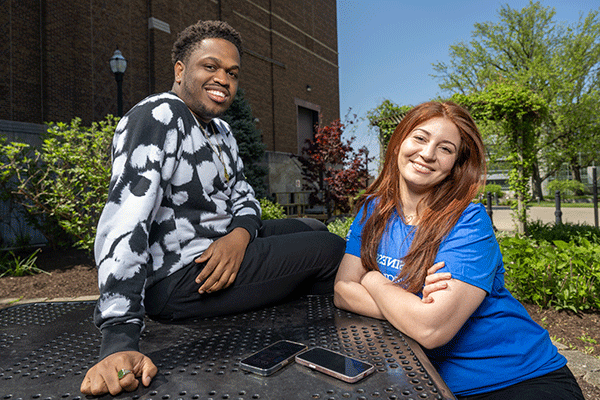 Two students, one female sitting at a table and one male sitting on the table, smile and pose